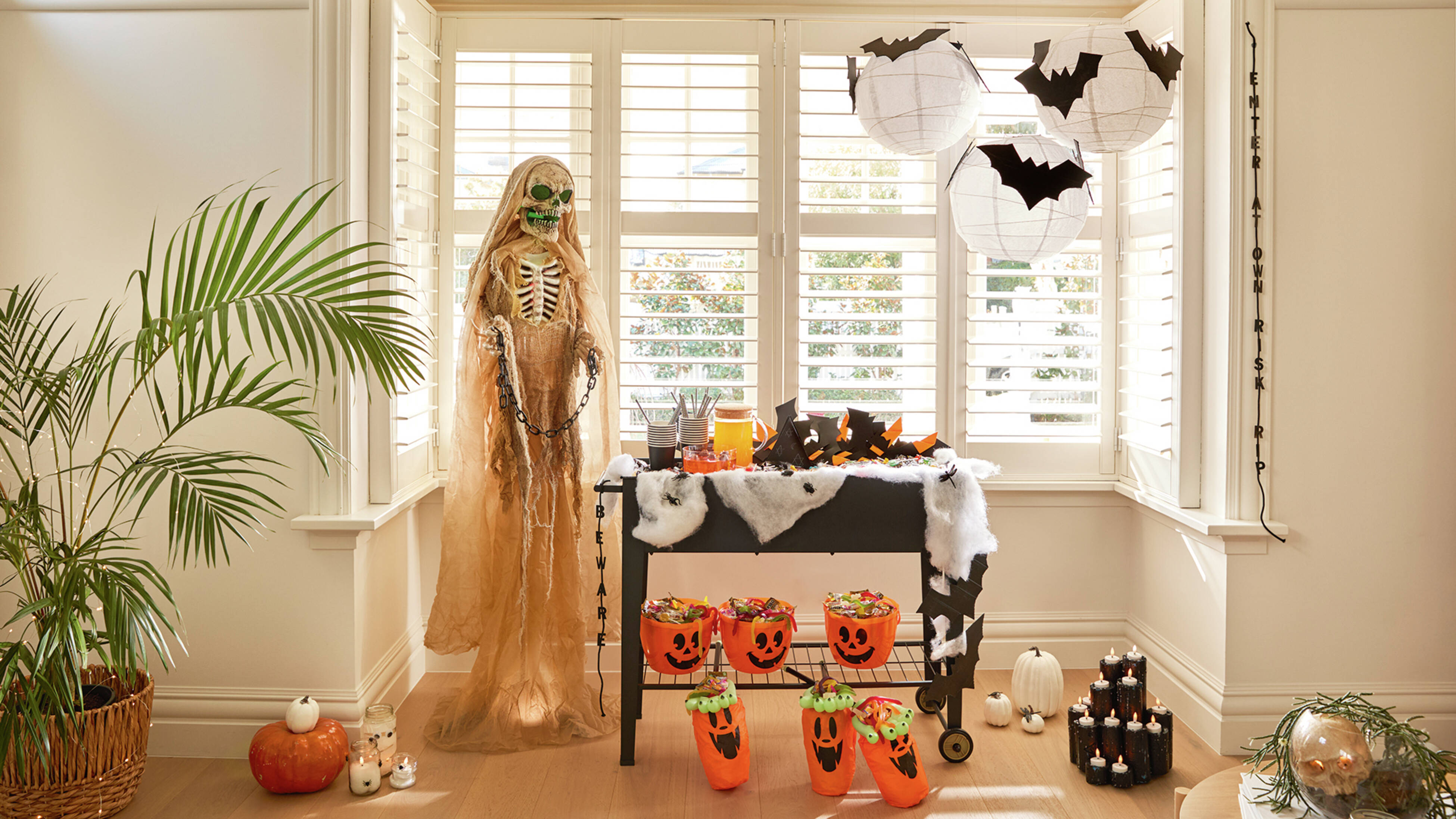 A bright corner decorated for Halloween with a standing skeleton, a table covered in spider webs and bat cutouts, hanging paper lanterns with bats, and orange pumpkin buckets filled with treats.