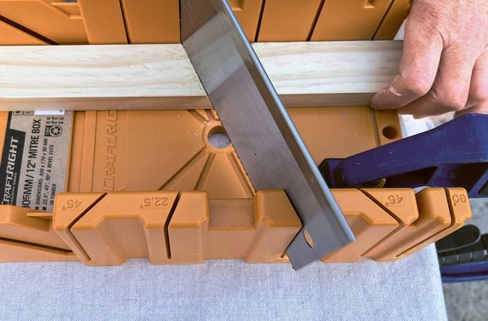 A hand holding a piece of pine in a yellow mitre box, with a saw cutting the timber.