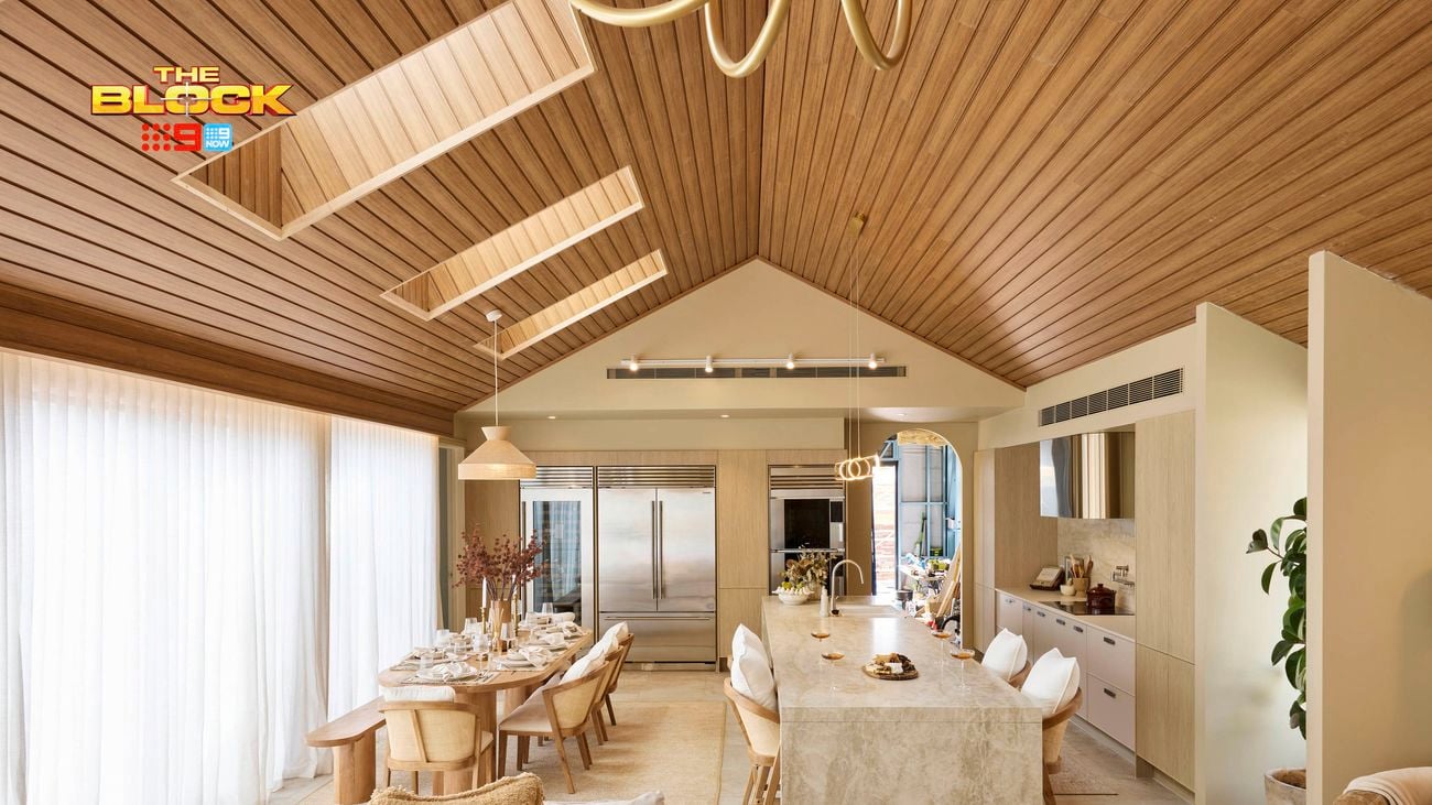 Open-plan kitchen and dining space from The Block, featuring a vaulted timber ceiling, marble island with bar stools, large fridge, neutral-toned dining area, and floor-to-ceiling curtains.