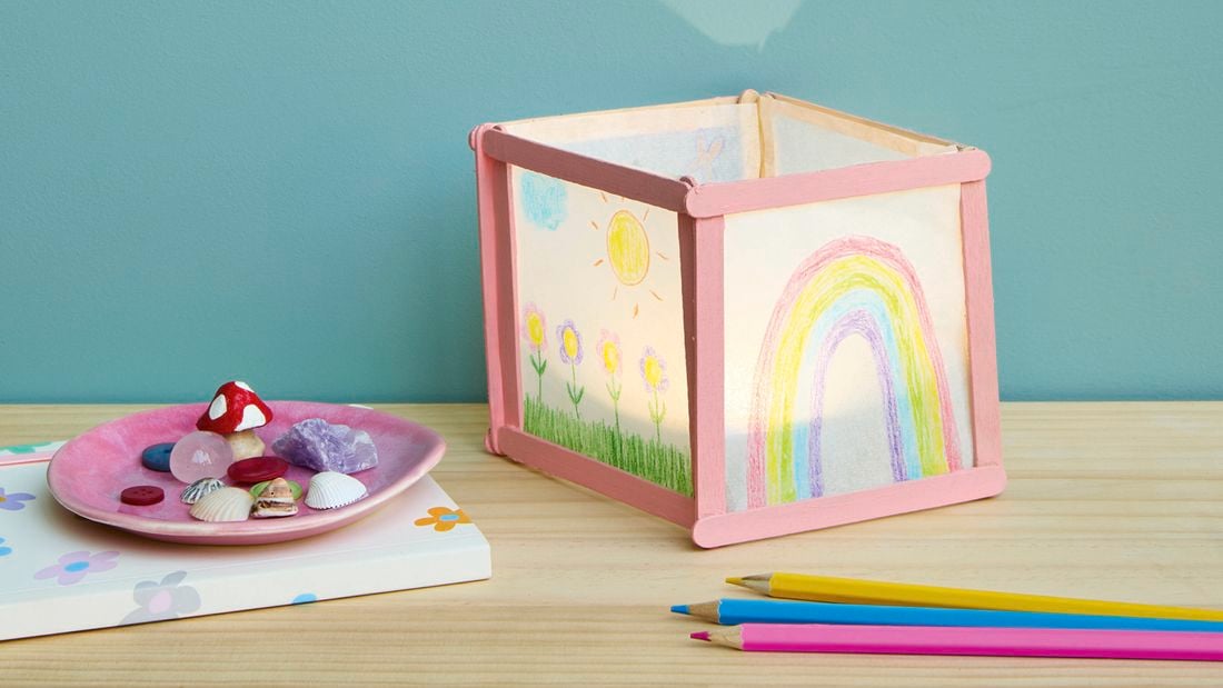 A handmade paper lantern decorated with children’s drawings of flowers, a sun, and a rainbow, sitting on a wooden table alongside coloured pencils and a small dish of shells and trinkets.