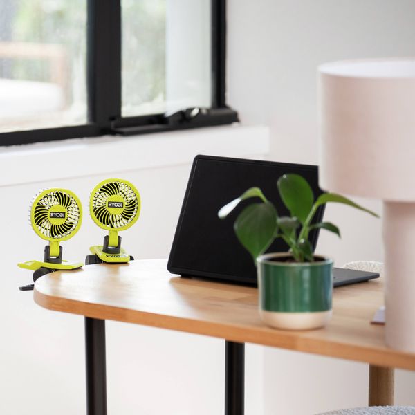 Two compact Ryobi clip-on fans attached to a desk beside a laptop and potted plant near a window.