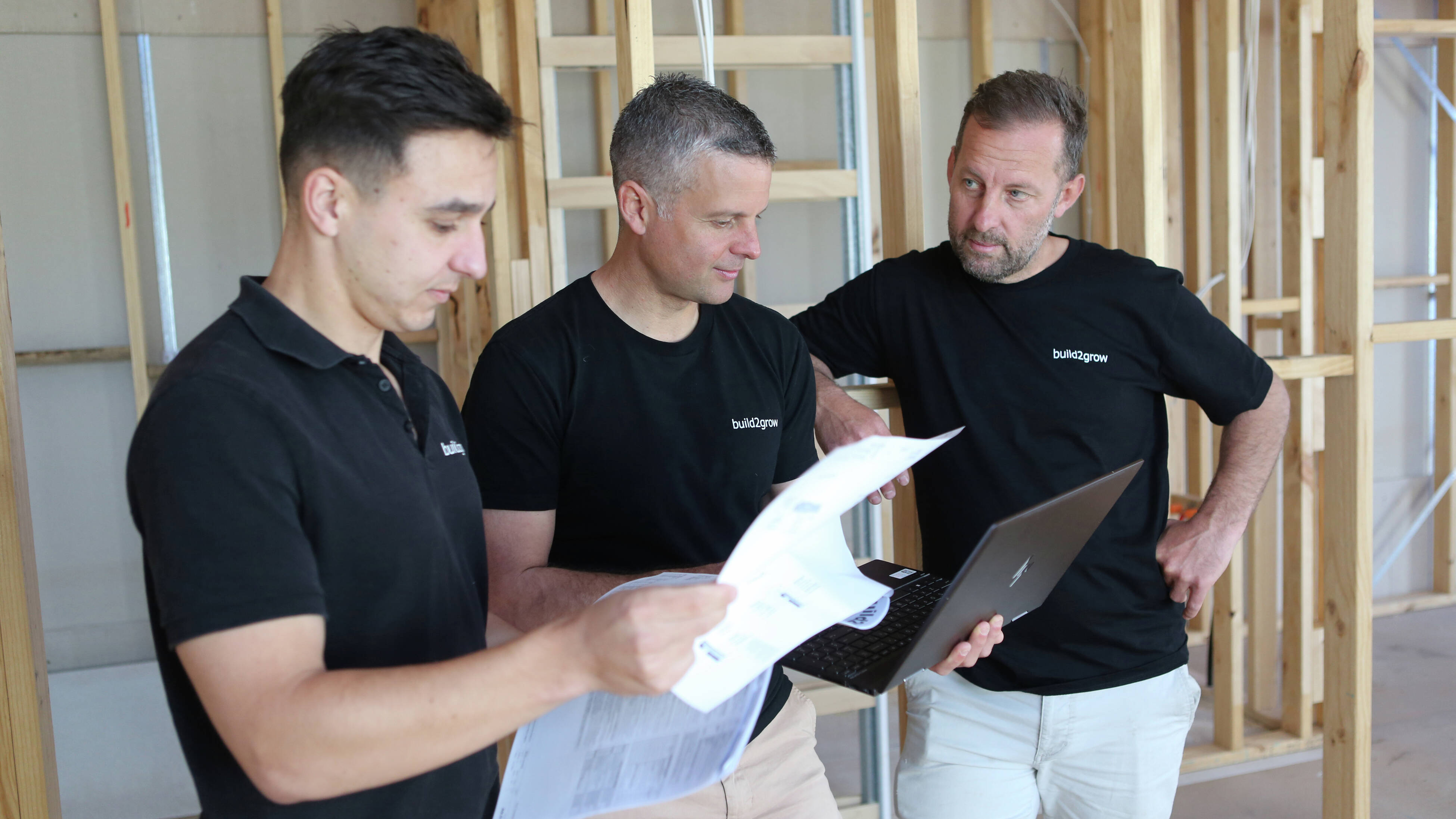 Director of build2grow Michael Nikolovski wearing a black shirt and beige pants, sits on scaffolding at a building site, holding an open laptop.]