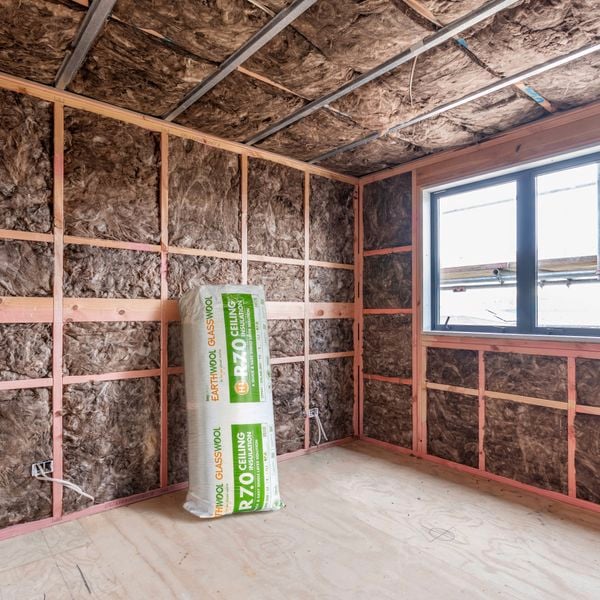 Unfinished room under construction with exposed timber framing and brown insulation batts lining the walls and ceiling, a sealed bag of R7.0 glass wool ceiling insulation standing on the wooden floor, and a rectangular window letting in daylight.