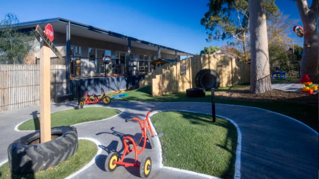 The outdoor playground of a kindergarten, with tricycle, concrete track, tanbark area and big tree, in front of a large building.