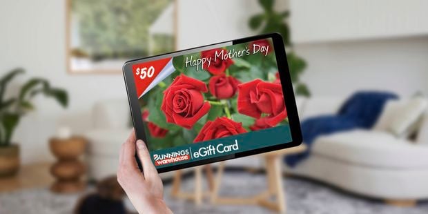 A person holding a tablet with a Bunnings $50 gift card with roses in the background on the screen in a lounge room.