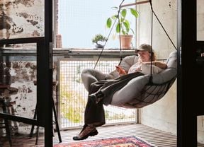 Cody Weightman relaxes in a hanging swing chair in front of a large window, surrounded by stone walls, timber flooring, and a red and blue rug