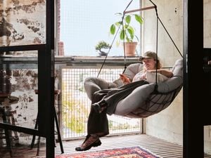 Cody Weightman relaxes in a hanging swing chair in front of a large window, surrounded by stone walls, timber flooring, and a red and blue rug