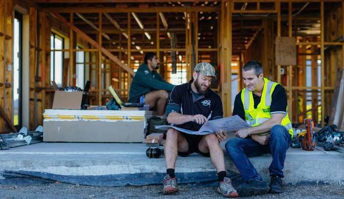 Two people wearing high‑visibility vests and hard hats stand at a construction site. One holds a rolled-up document. Behind them are construction materials, a partially framed structure, and an excavator.