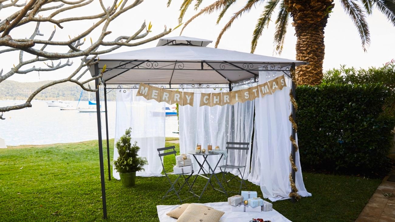 A white gazebo with a black metal frame installed on a lawn outside, with a table and chairs, picnic rug with cushions, and Christmas decor.