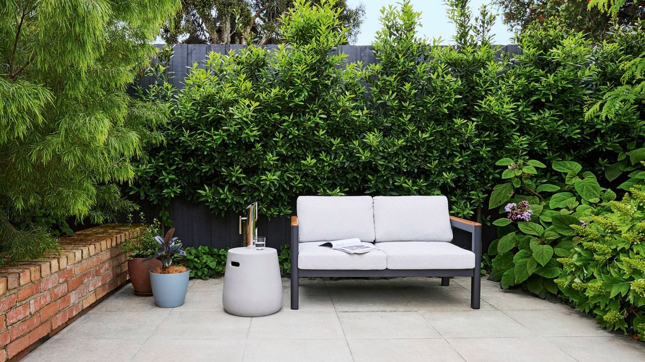 A courtyard with large light-coloured paving, a brick retaining wall to the left, a dark grey timber fence to the back and plantings on all three sides. A two seater outdoor sofa sits next to a concrete look side table and two pot plants.