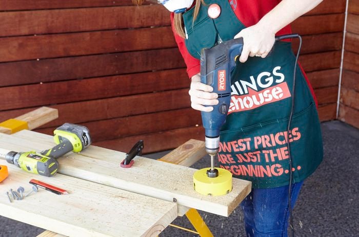A person cutting a large hole in a pine board using a hole saw attachment on a drill