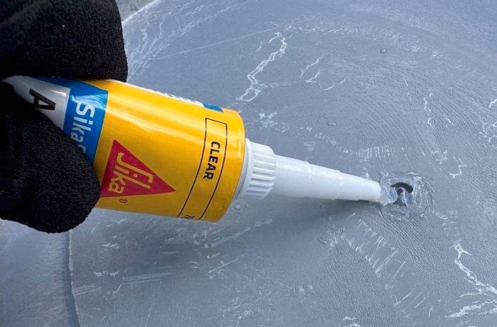 A gloved hand applying clear silicone sealant from a tube onto a small hole in a grey surface.   