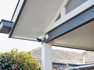 White security camera installed under a house roof eave above an outdoor area, with shrubs and neighbouring rooftops visible in the background.