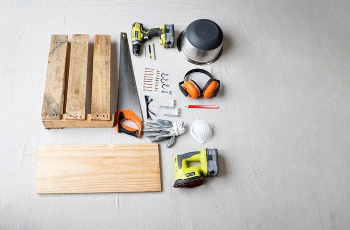 Timber, saw, drill, pencil, earmuffs, pot and more laid out on the ground.