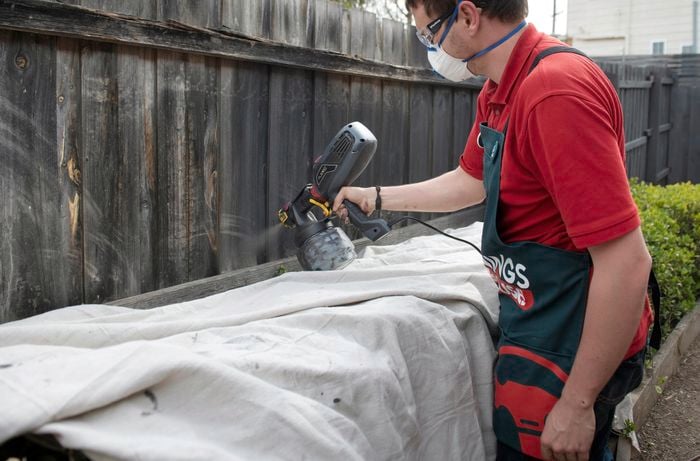 Person wearing a mask and safety glasses spray painting a paling fence