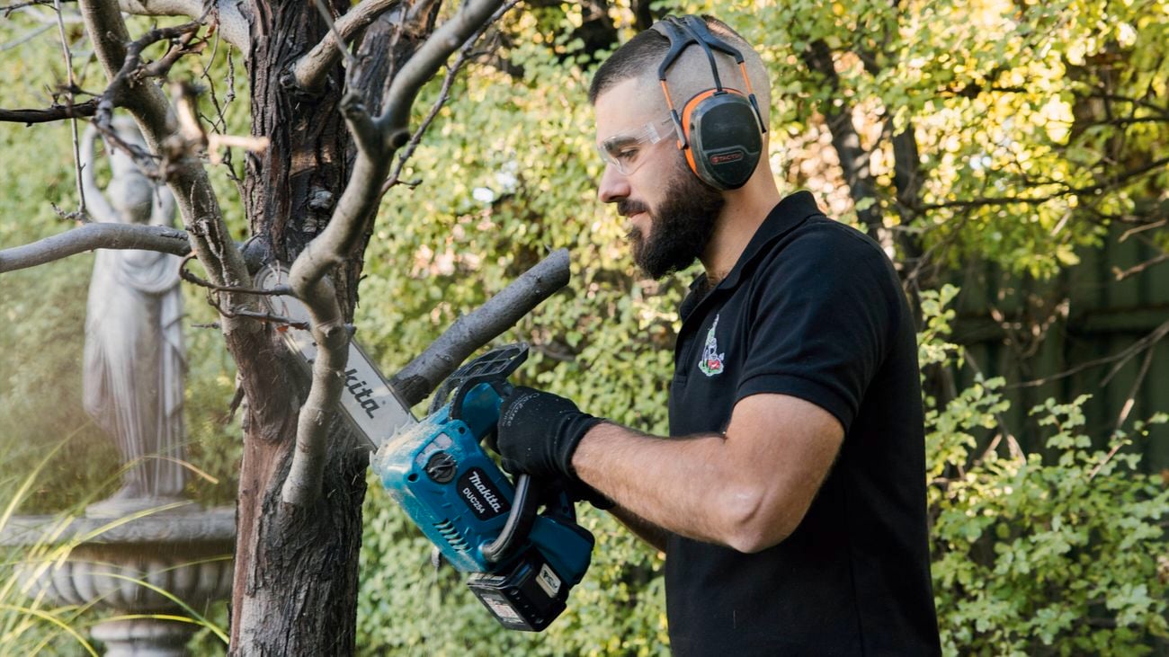 A man in a black shirt, ear muffs and gloves uses a Makita 18V 250mm chain saw to cut through a tree branch