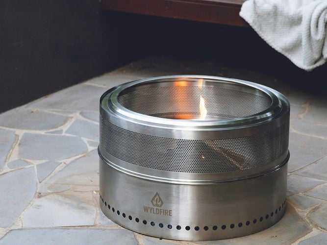 Stainless steel Wildfire fire pit lit with a small flame on a tiled patio, with a bench, towels, and wine bottles in the background.