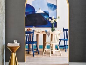 Grey wall with arched doorway, opening out into a white room with a blue abstract painting and a wooden dining table with blue and white chairs.