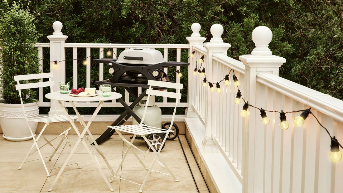 Deck with white balustrade, string lights, a gas barbecue, and a small outdoor dining set surrounded by greenery.