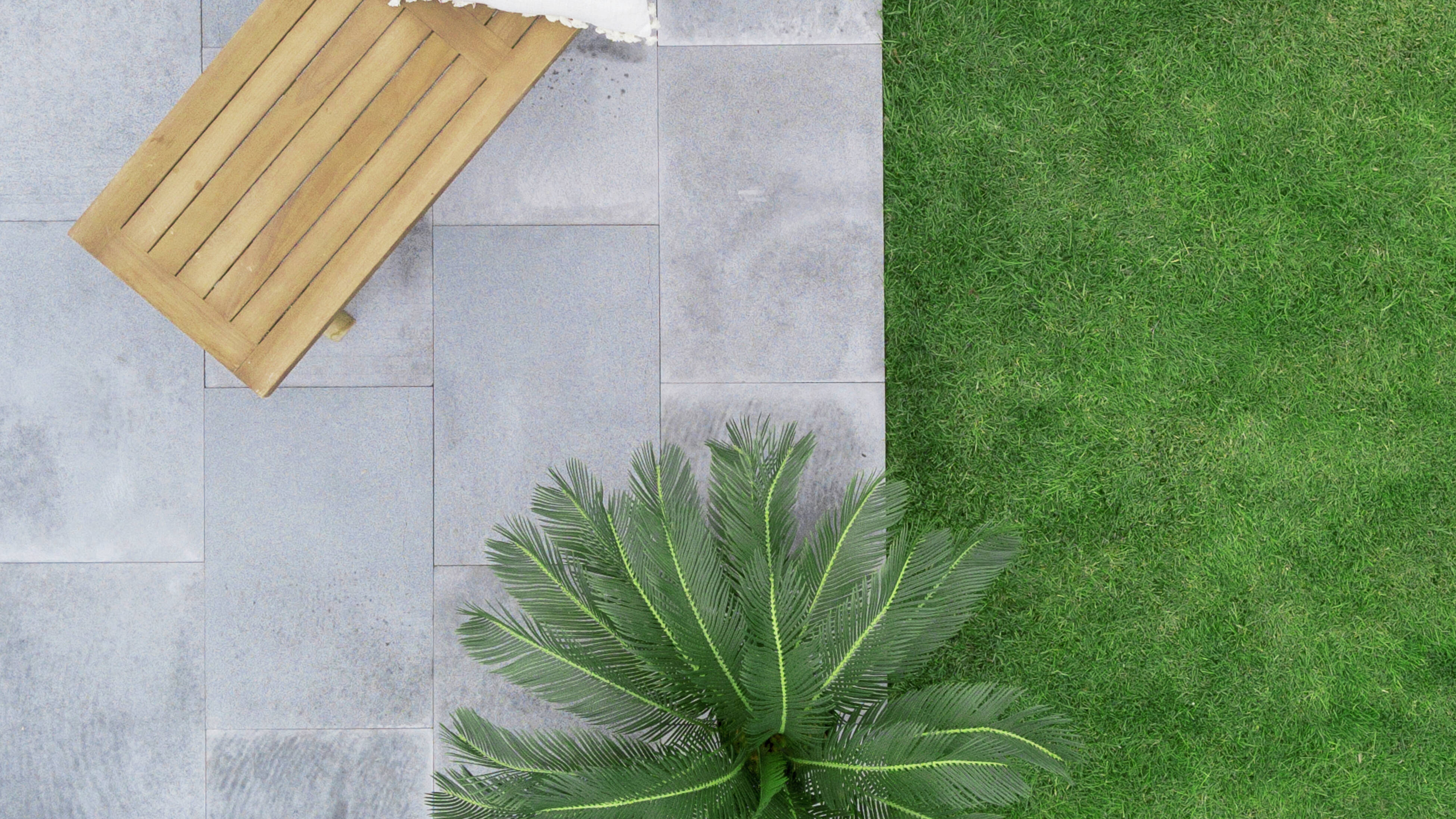 Top-down view of a grey stone larged paved patio with a wooden bench and white cushion, next to a green lawn and a palm plant