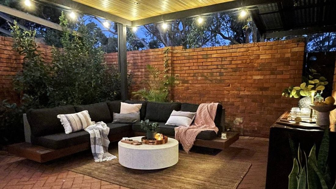 Outdoor timber floating lounge with grey cushions, throw rug, coffee table sitting in front with red brick wall in back ground
