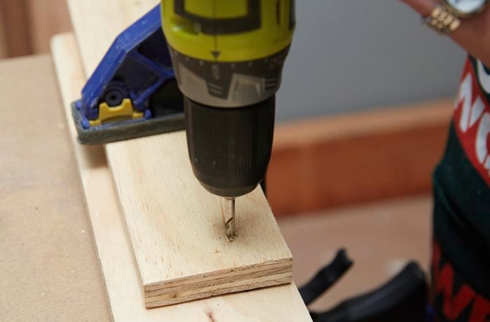 A person drilling through a piece of plywood clamped to another piece of wood