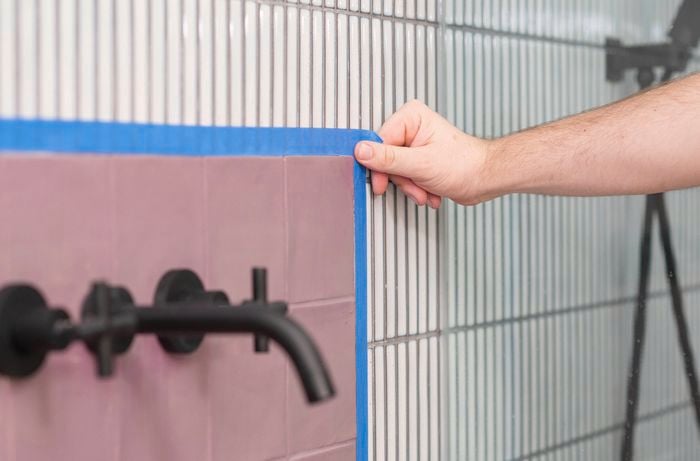 A person is applying painter's tape along the edges of tiles to cover those areas that don't need paint. 