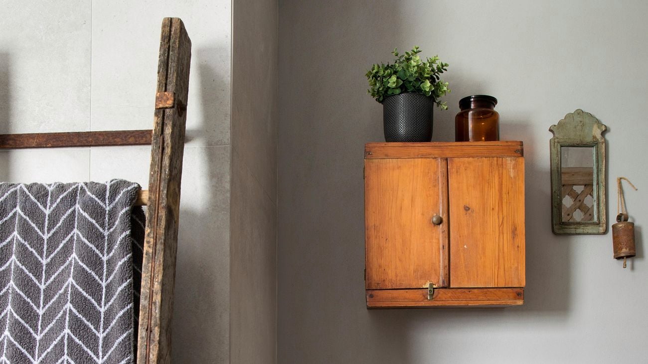 Bathroom with wooden towel rack, woven clothes basket and a small wooden cabinet attached to the wall with a pot plant on top.