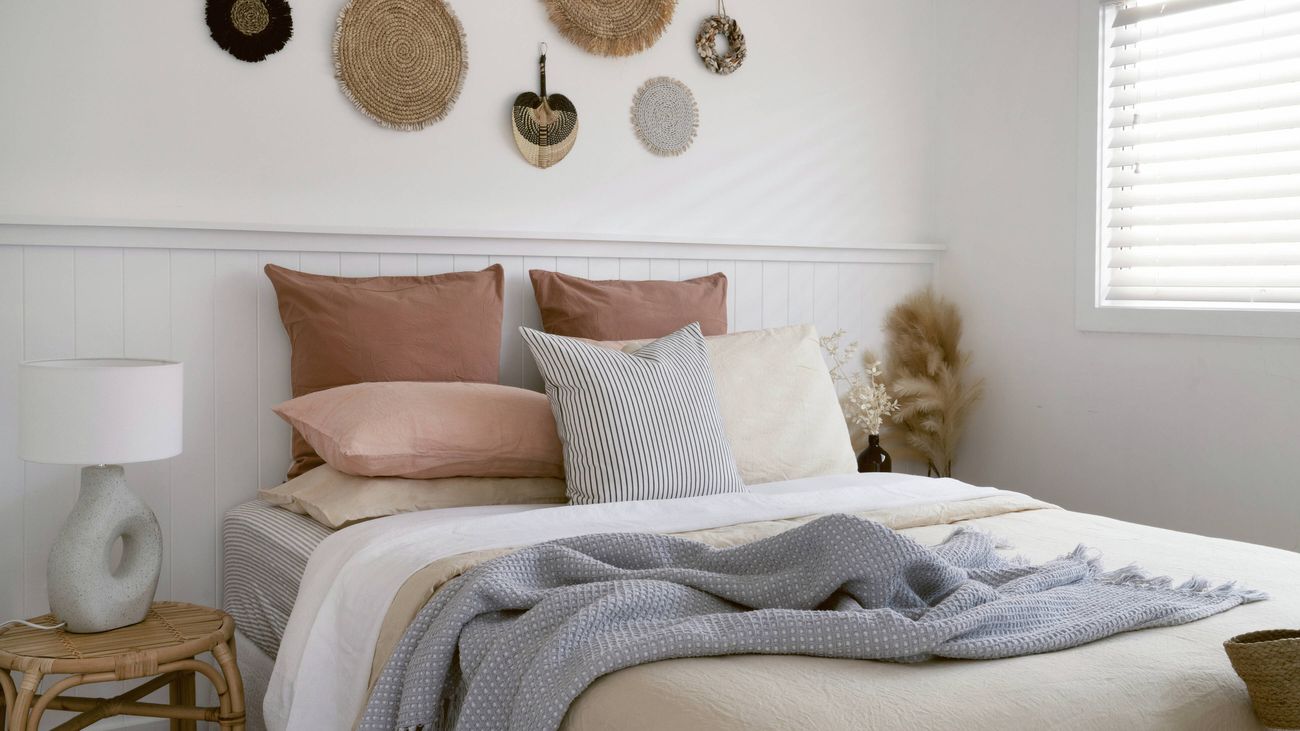 A bedroom with a bed featuring layered bedding in neutral tones, a rattan bedside table with a lamp, and a white dado wall decorated with woven wall hangings above it.