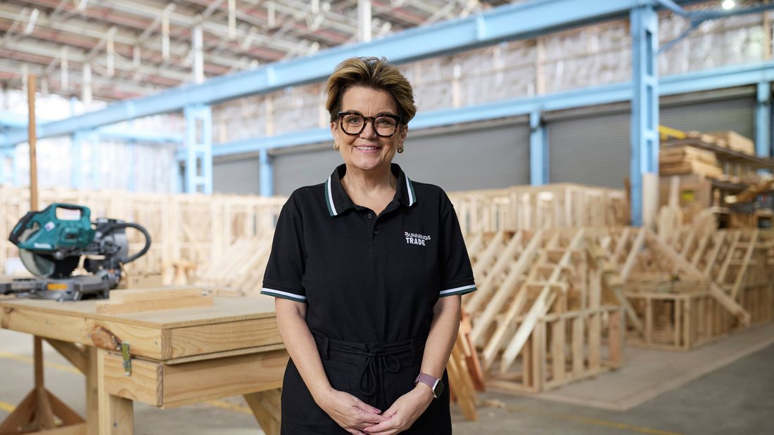 Bunnings Trade team member standing in a timber construction training workshop with woodworking equipment and timber framing in the background.