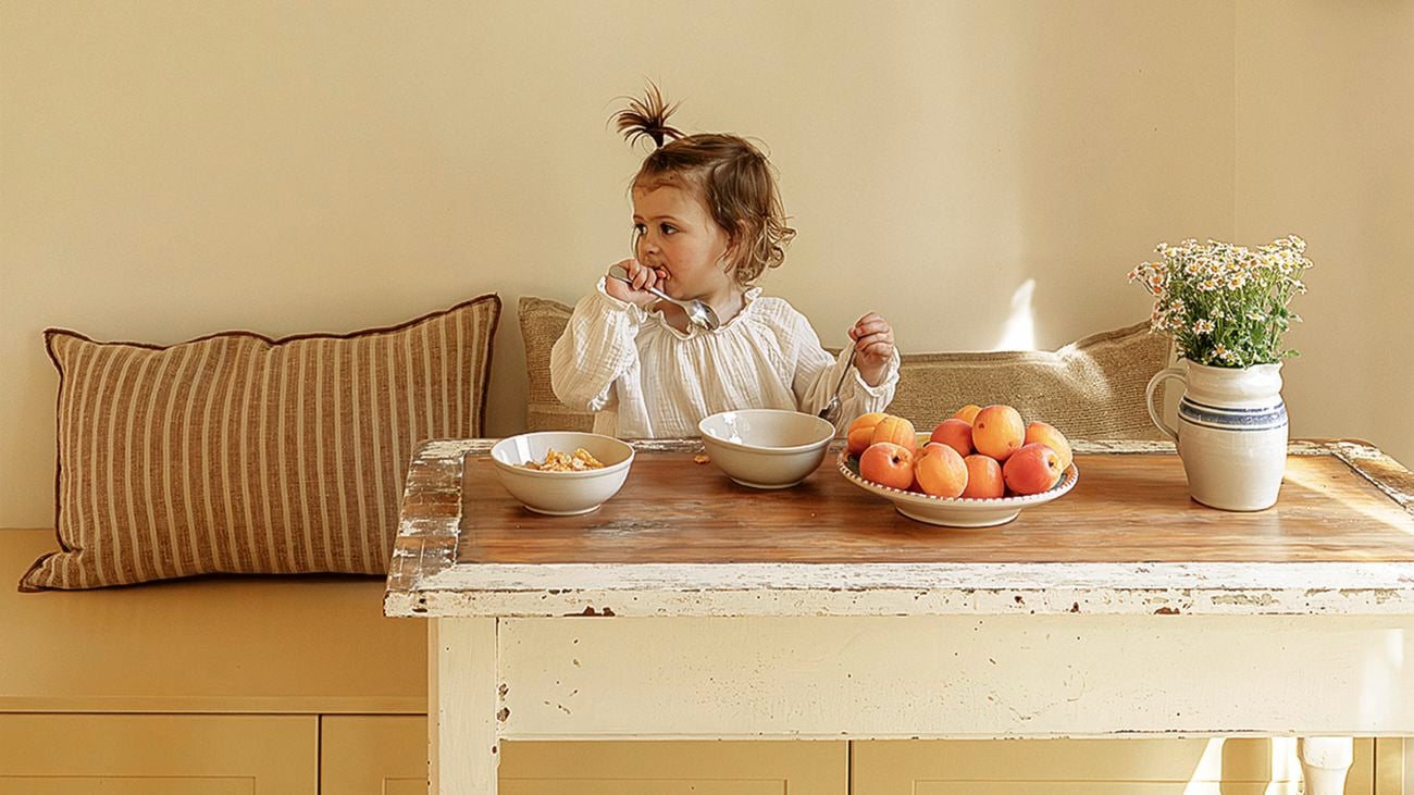 A toddler sits at a rustic wooden table in a breakfast nook with a built‑in yellow bench and striped cushion, eating from a bowl. On the table are a bowl of cereal, a plate of peaches and a vase of small white flowers.