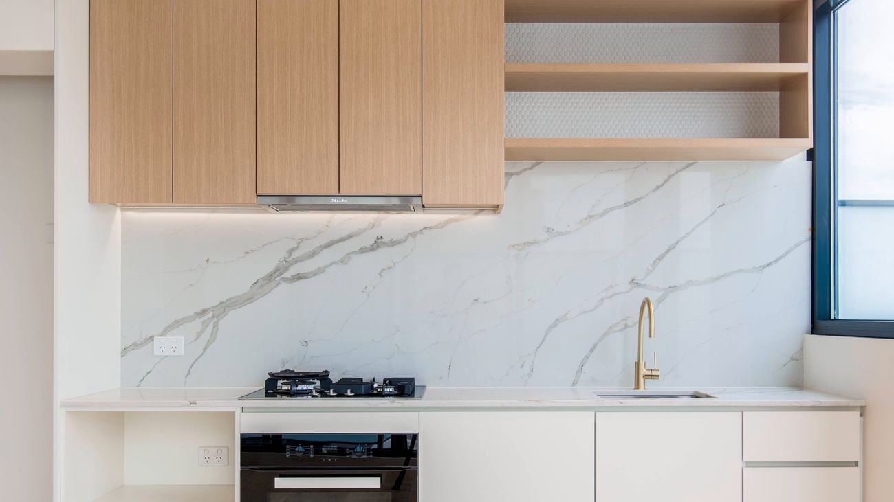 Modern kitchen with a white marble‑look splashback, timber upper cabinetry, white benchtops, a built‑in oven, gas cooktop and a gold mixer tap.
