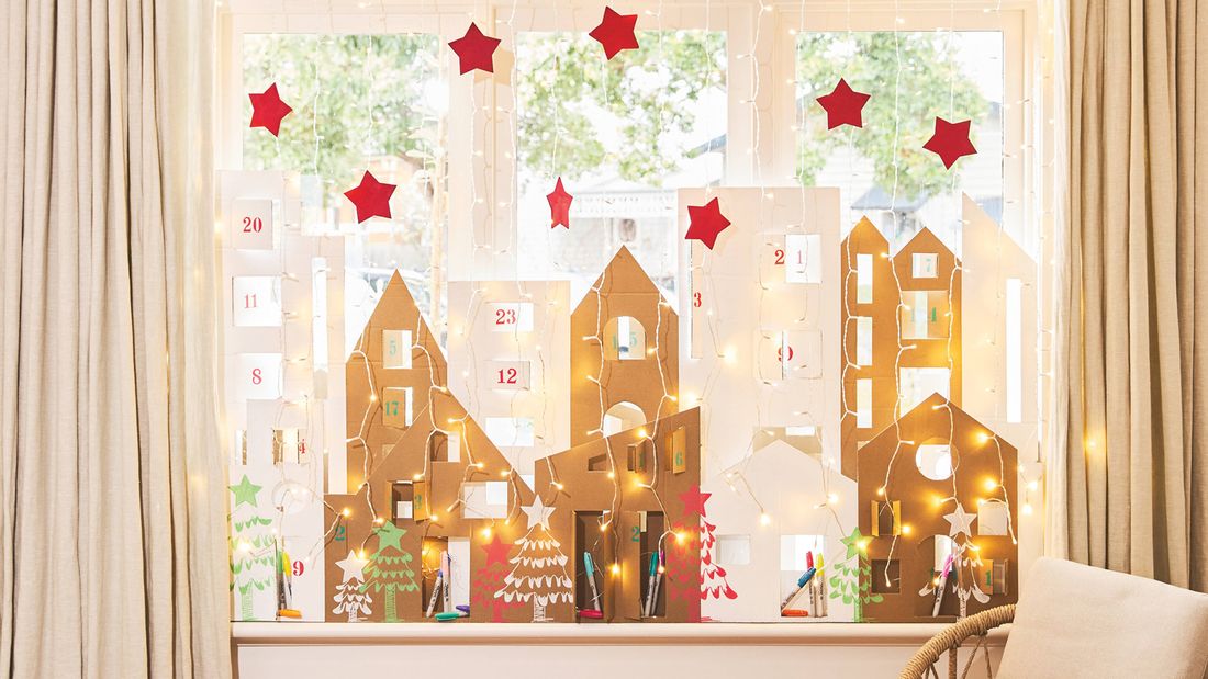 A Christmas window display featuring a homemade cardboard Advent calendar village on the sill, framed by waterfall lights and red craft stars.]