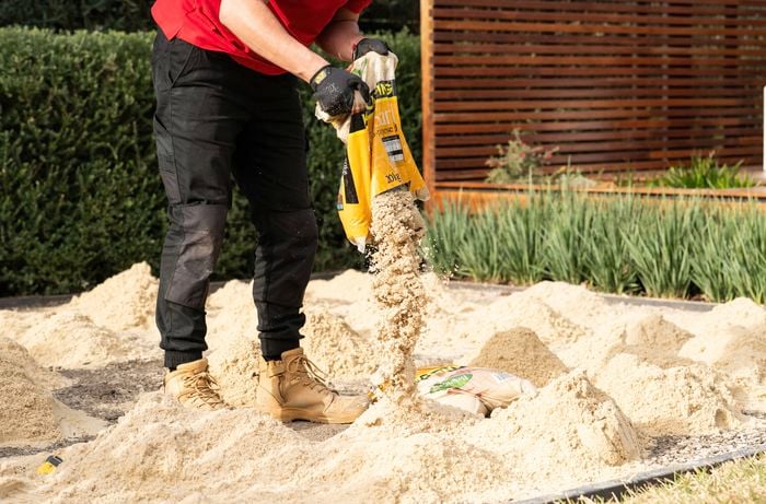A person wearing gloves and work boots is spreading paving sand from a bag onto the prepared ground surface.