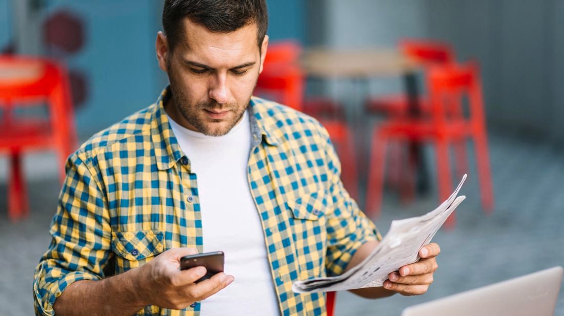 A man in yellow and blue checked shirt looks down at his phone as he holds a newspaper