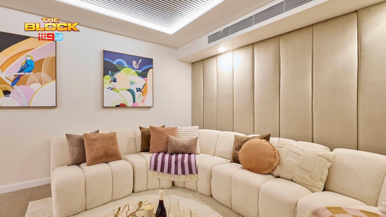 A cozy living room with a beige sectional sofa decorated with assorted throw pillows and blankets. A round coffee table in the center holds popcorn, books, and bottles. The walls feature two colorful abstract paintings, and the ceiling has recessed lighting, contributing to the room’s modern and inviting atmosphere.