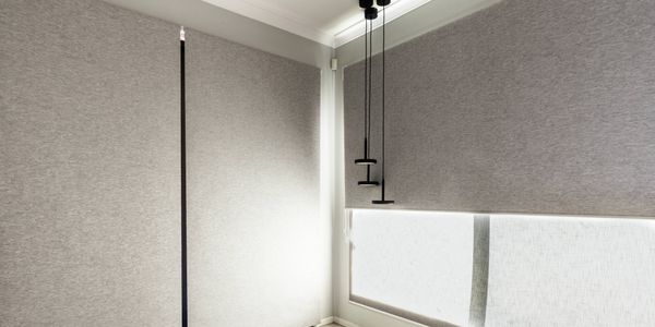 Light grey roller blinds covering large corner windows in a modern living area, with black pendant lights suspended in front of the windows and soft natural light filtering through the fabric.
