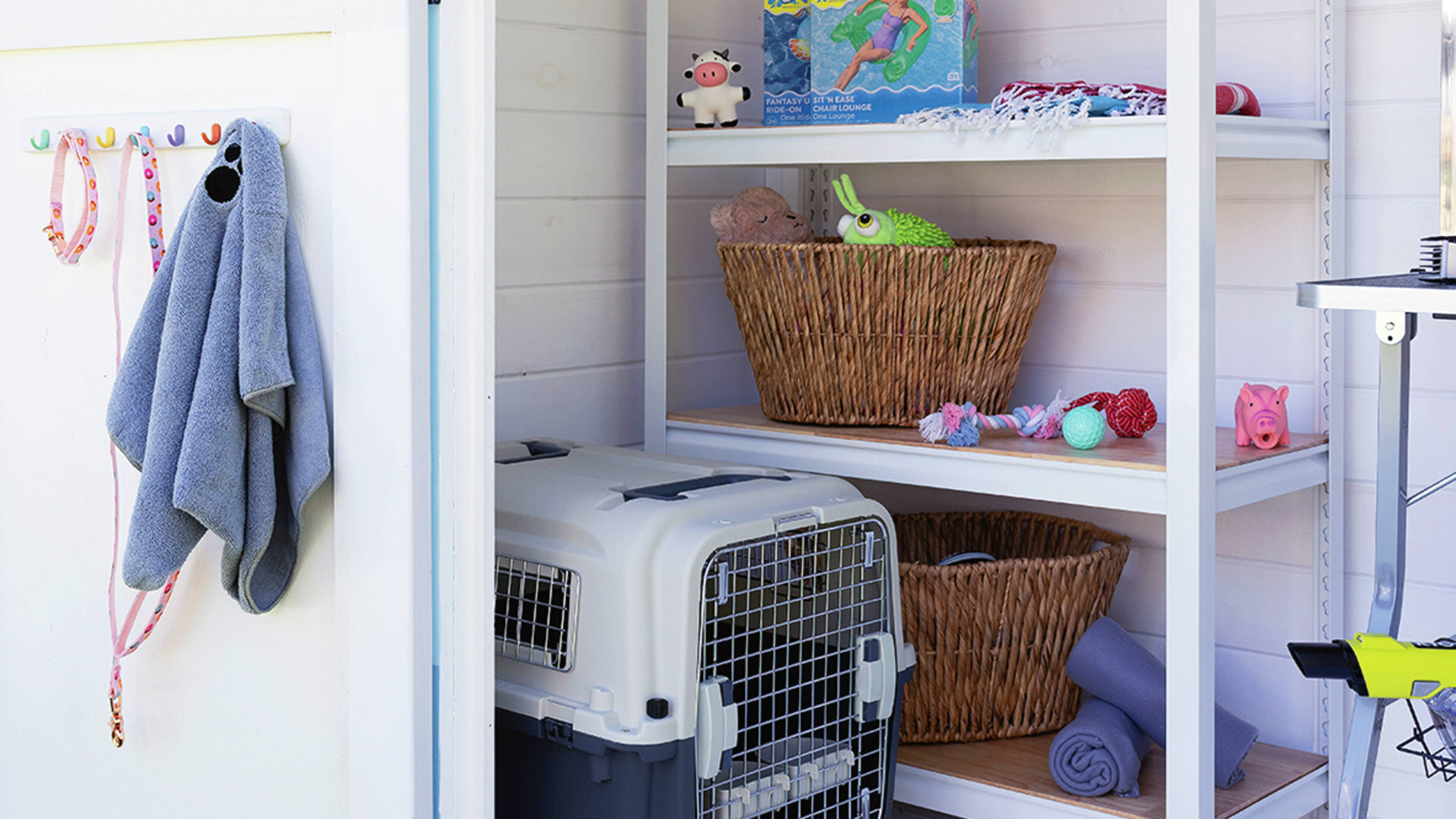  Inside a small shed, white shelves hold woven baskets, pet toys, towels, and grooming bottles. A grey pet carrier sits on the floor with a pink bowl in front