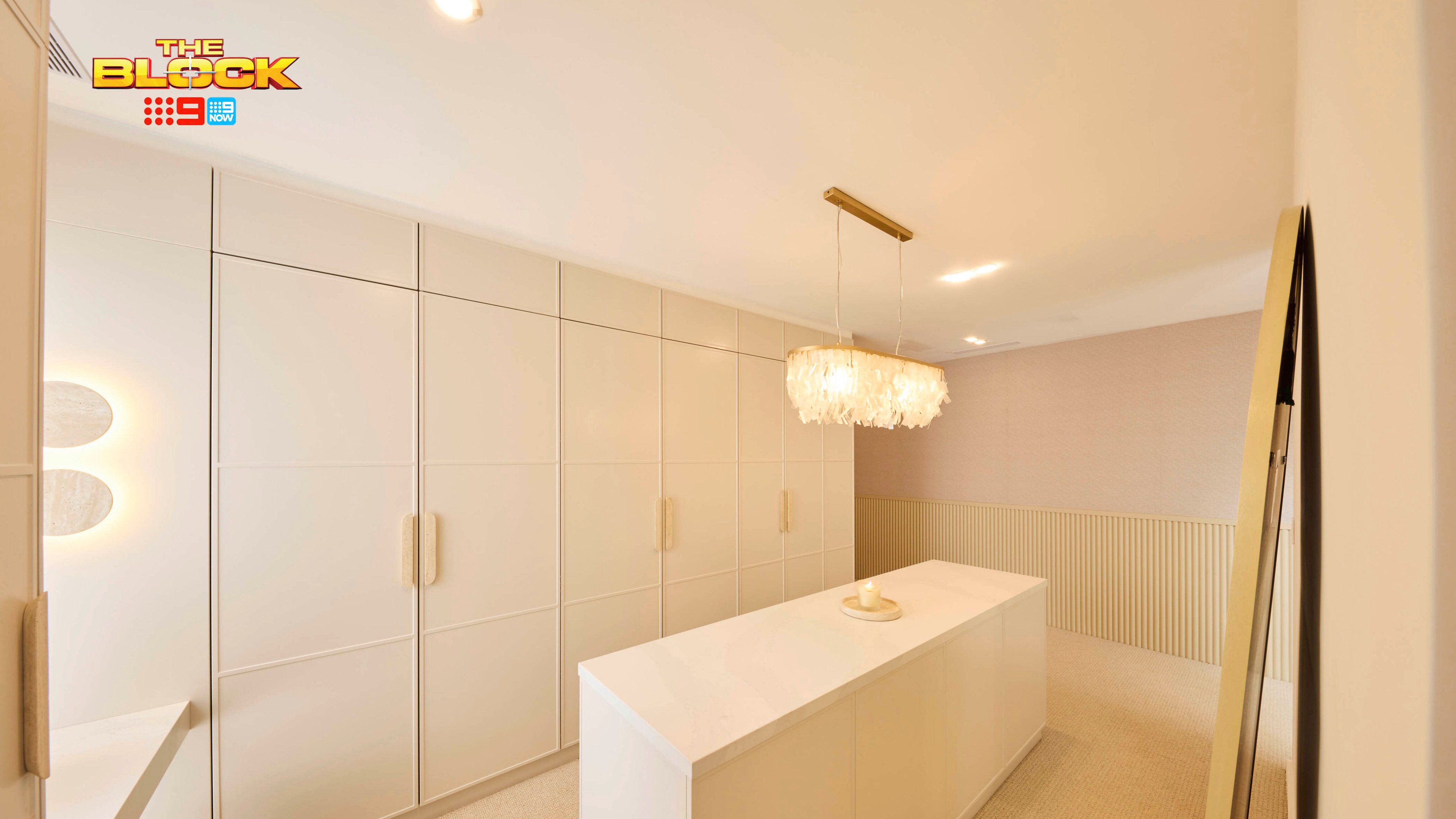 A modern walk-in wardrobe with a central island topped with a white countertop and a decorative item. Built-in wardrobes with sleek, handle-less doors line one wall. A stylish chandelier hangs above the island, and one wall features textured paneling. The space is painted in neutral tones and well-lit. 