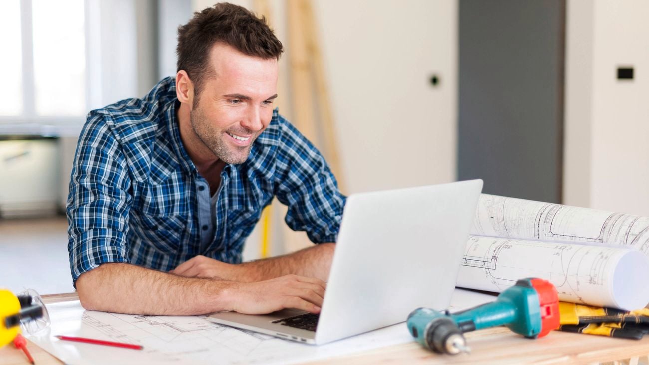 A man in a blue plaid shirt smiles while working on a laptop, with architectural plans and tools on the desk.