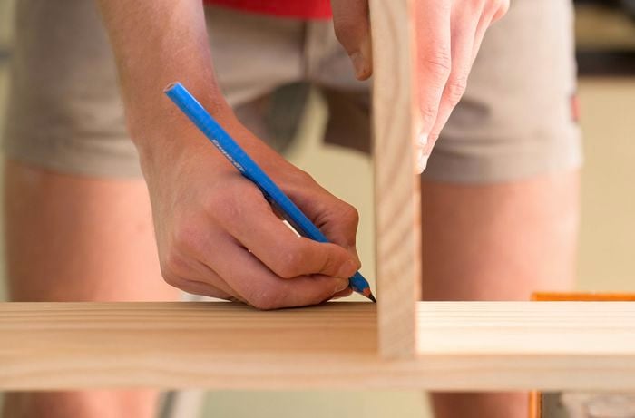  A person is using a vertical board as a guide to mark the joint position on a wooden bookshelf panel with a pencil