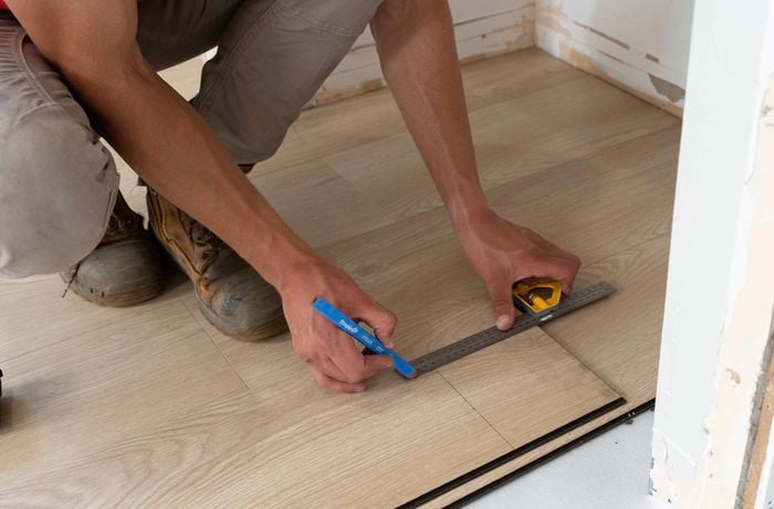 A person is measuring and marking a laminate floorboard using a combination square and a blue pencil to work around corners.