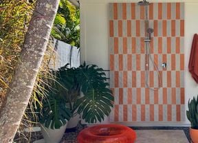 Outdoor shower with orange and white tile panel, overhead shower and hand shower. An orange towel, plants and pool toys sit nearby.