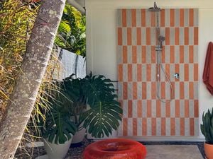 Outdoor shower with orange and white tile panel, overhead shower and hand shower. An orange towel, plants and pool toys sit nearby.