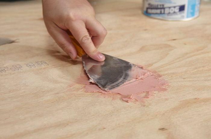 A person filling a hole in plywood using builders bog and a metal scraper