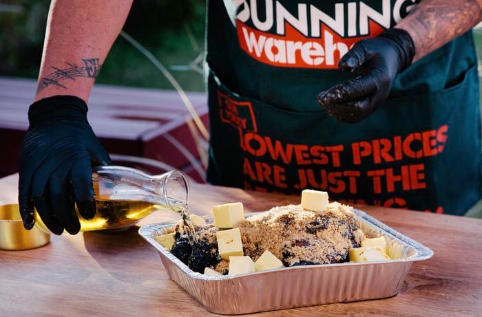 Person pouring apple juice over pork shoulder with butter cubes in cooking tray