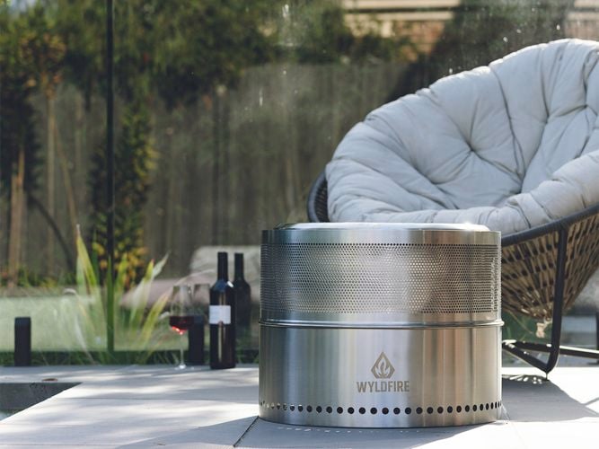 Compact stainless steel Wildfire fire pit placed on a patio beside lounge seating and wine bottles in a modern outdoor setting.
