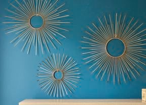 Three gold sunburst wall art pieces made from cable ties arranged on a teal wall, each featuring a circular center with evenly spaced radiating spokes in varying sizes, displayed above a light wooden surface with a woven storage box.