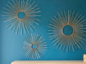 Three gold sunburst wall art pieces made from cable ties arranged on a teal wall, each featuring a circular center with evenly spaced radiating spokes in varying sizes, displayed above a light wooden surface with a woven storage box.