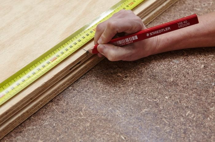 A person using a measuring tape to mark a measurement on a sheet of plywood
